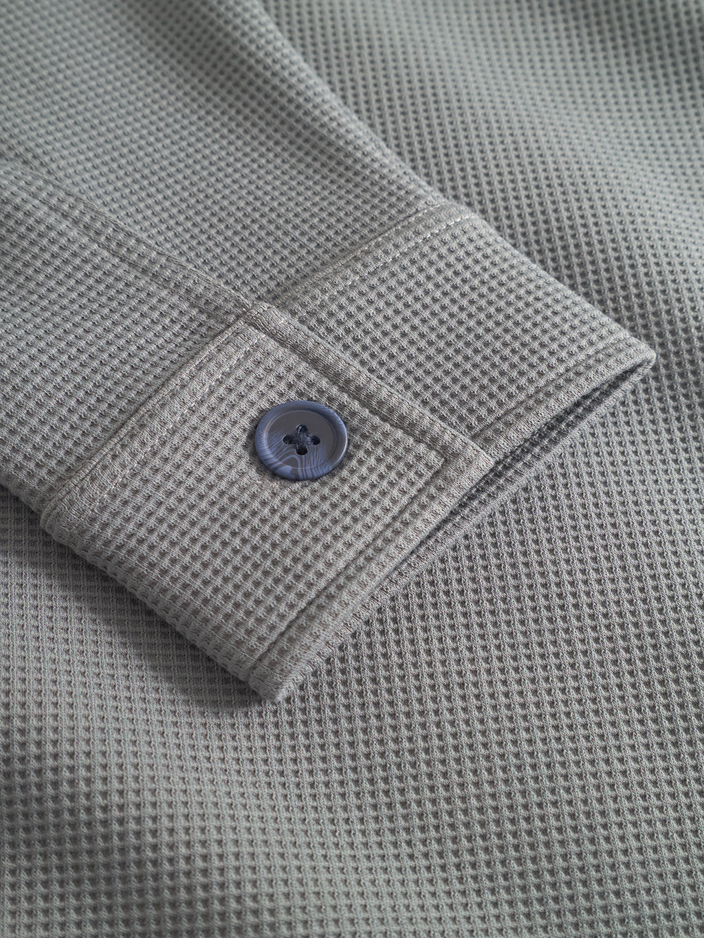 A close-up of the Como Waff - Dust Sage Green overshirt by Gabba shows its waffle-knit, grid-patterned sleeve with a cuff and dark gray button—perfect for stylish, warm layering.