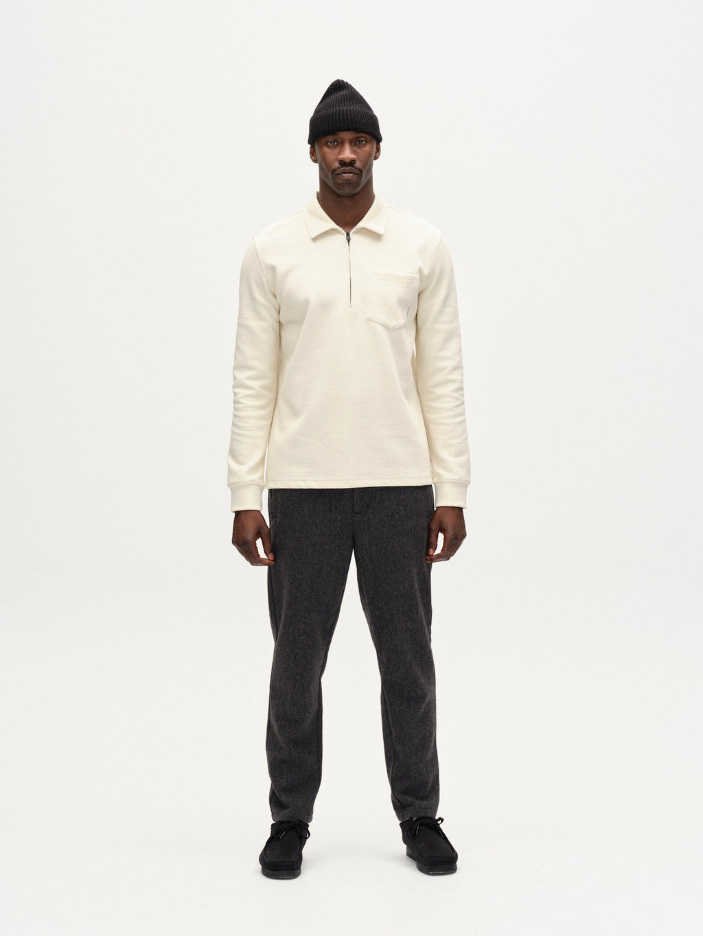 A man faces forward against a plain white background, wearing the Gabba Island Waff shirt in White Almond, dark gray pants, black shoes, and a black beanie.