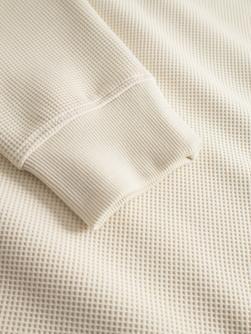 A close-up of the Gabba Island Waff – White Almond shirt showcases mens fashion with its cream waffle-knit texture, long sleeves, and ribbed cuffs, laid flat on matching fabric.