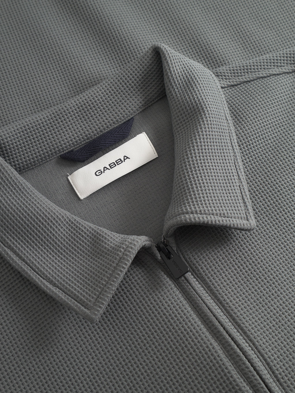 Close-up of the Island Waff - Dust Sage Green zip-up jacket by Gabba, featuring a textured waffle surface, pointed collar, and an inner neckline label with the brand name.