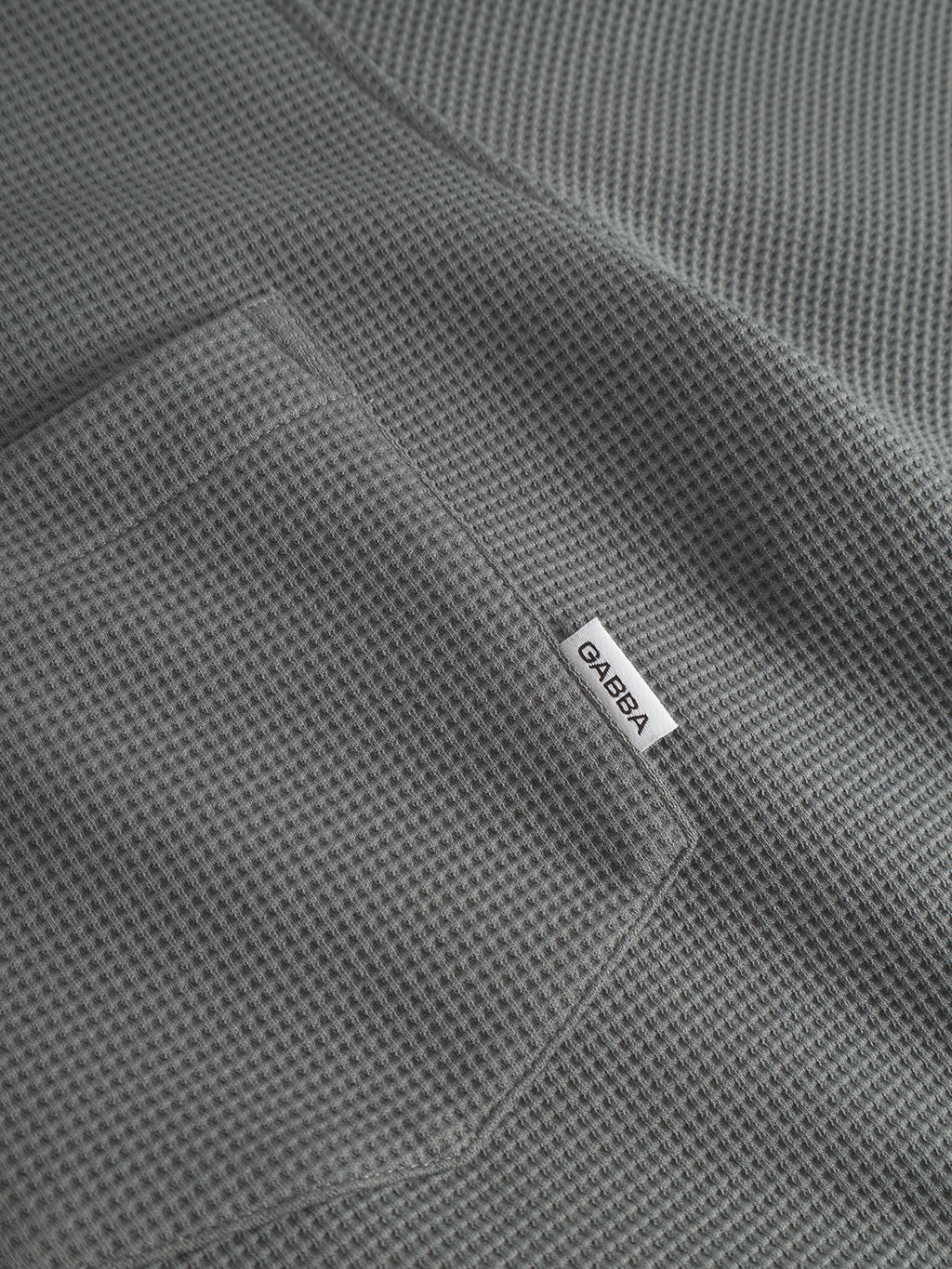Close-up of Island Waff - Dust Sage Green by Gabba: textured waffle surface, sewn-on pocket, and a small white tag reading GABBA in black on the pocket seam.