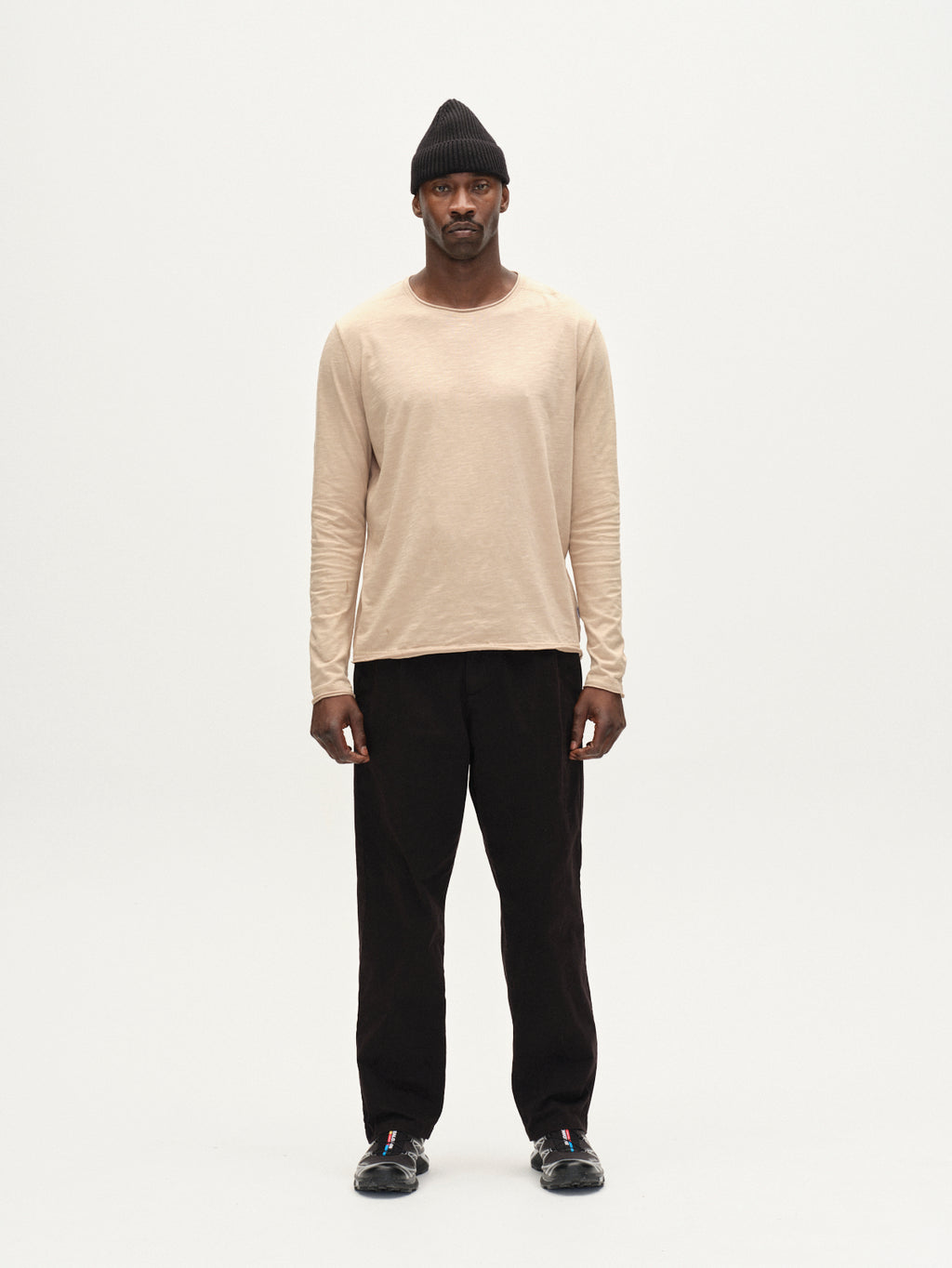 A man faces forward against a white background, wearing Gabbas Kostant New LS - Humus long-sleeved t-shirt, paired with black pants, black shoes, and a black beanie.