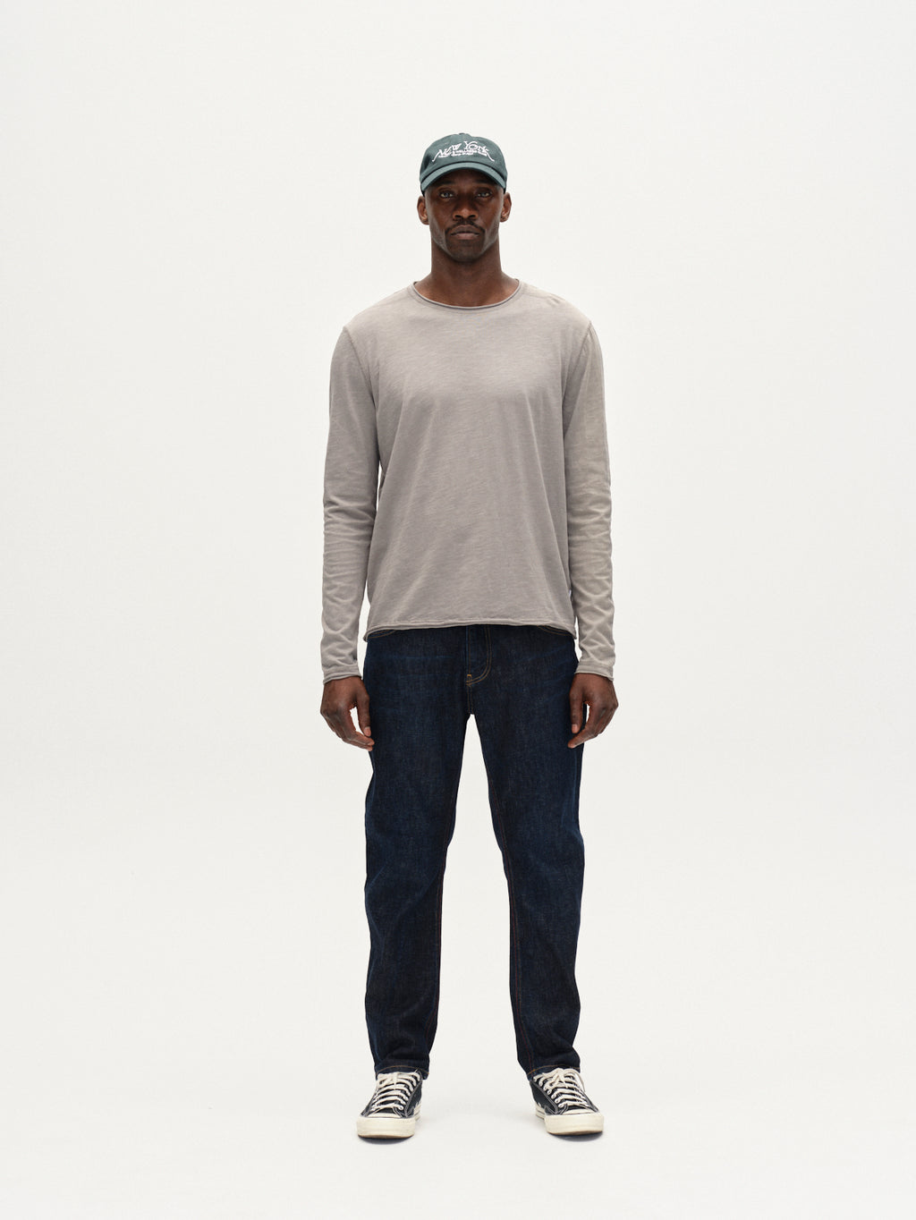 A man stands facing forward against a plain white background, wearing Gabbas Kostant New LS long-sleeved t-shirt in Dust Sage Green, paired with dark blue jeans and black-and-white sneakers.