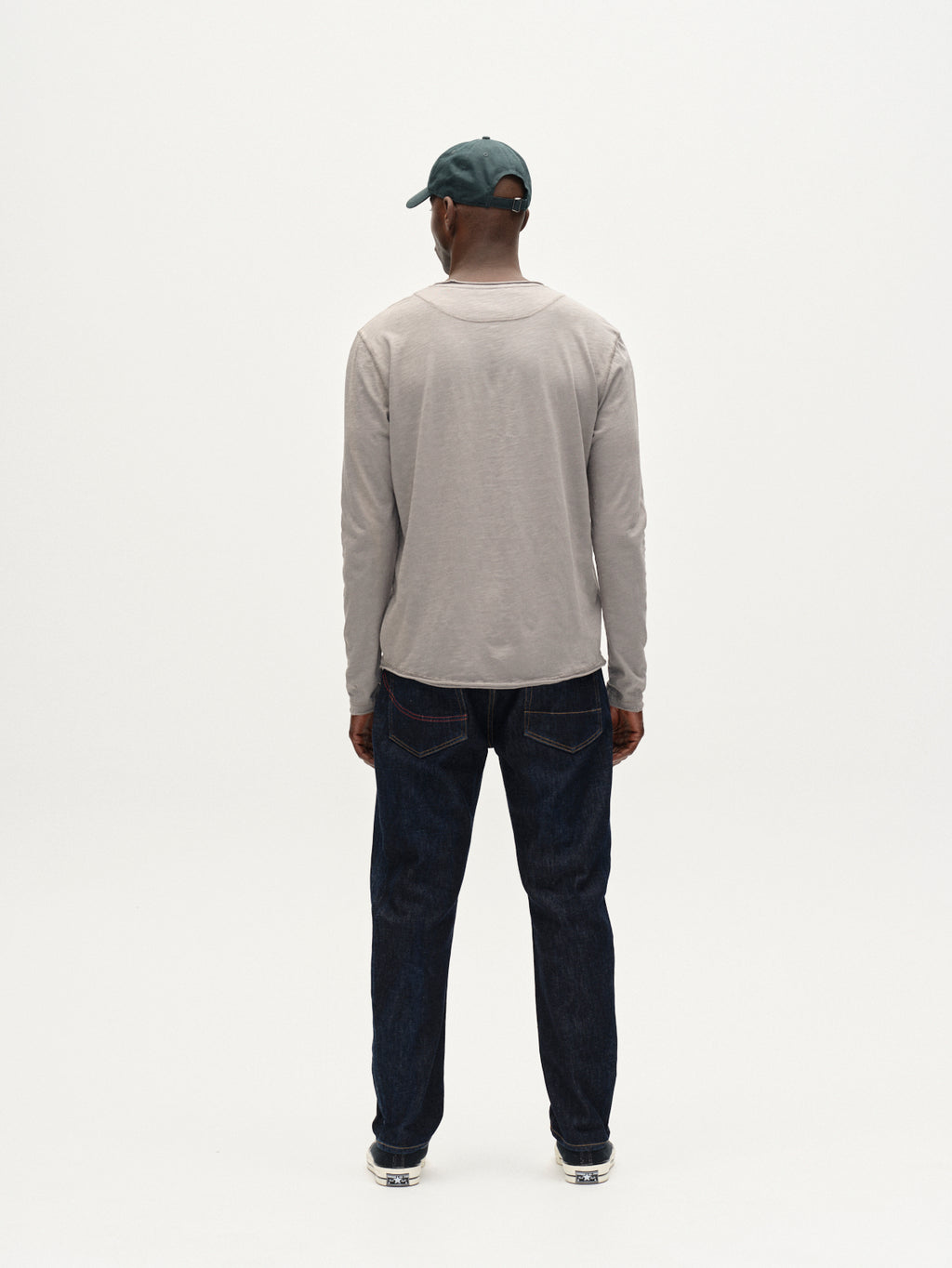 A man faces away from the camera, wearing the Gabba Kostant New LS in Dust Sage Green, a midweight slub jersey long-sleeve tee with raw edges, paired with dark blue jeans and silver sneakers against a plain white background.