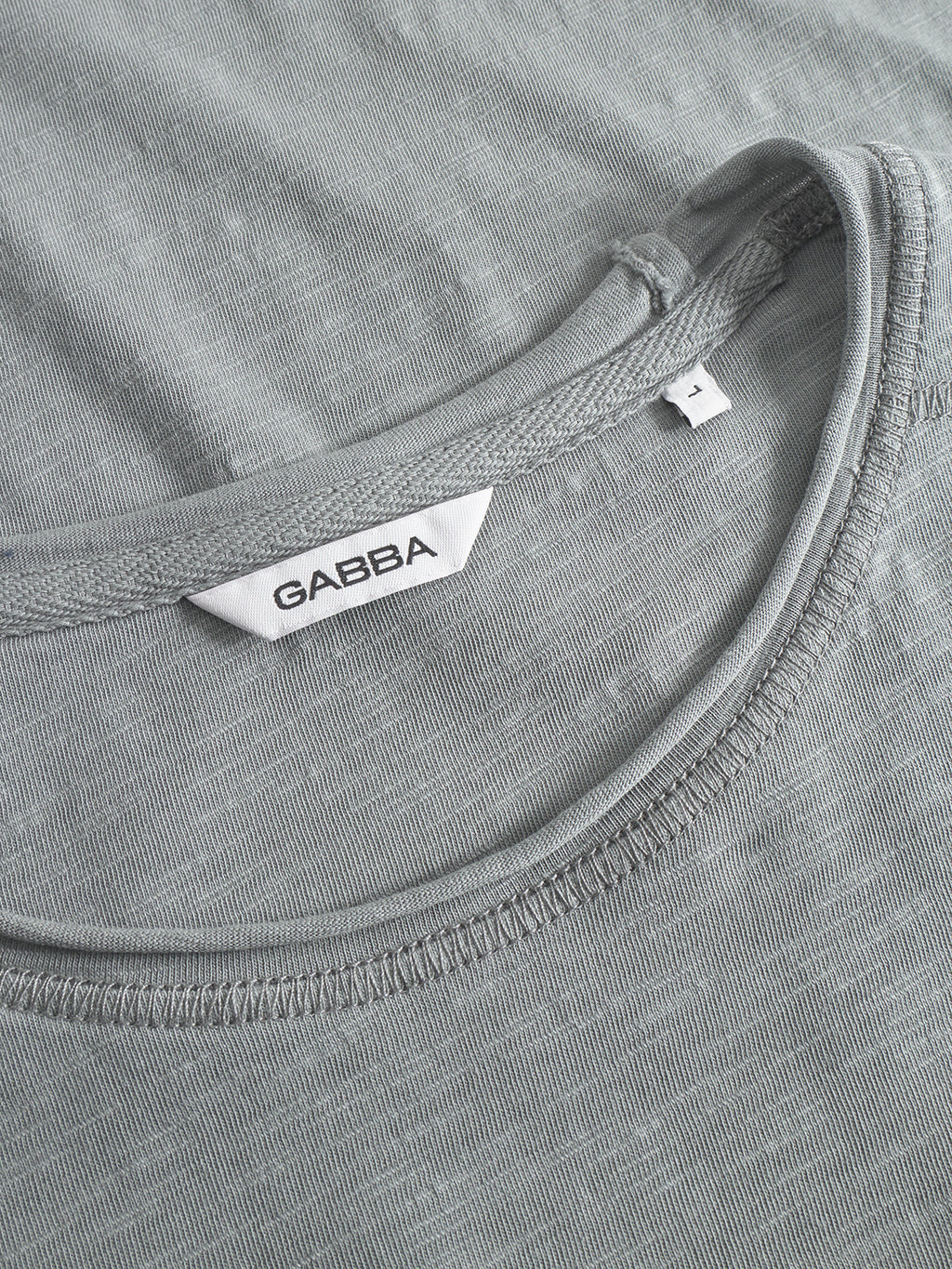 Close-up of the Kostant New LS T-shirt in dust sage green by Gabba, midweight slub jersey with a round neckline, featuring a white Gabba label and a small tag marked size M.