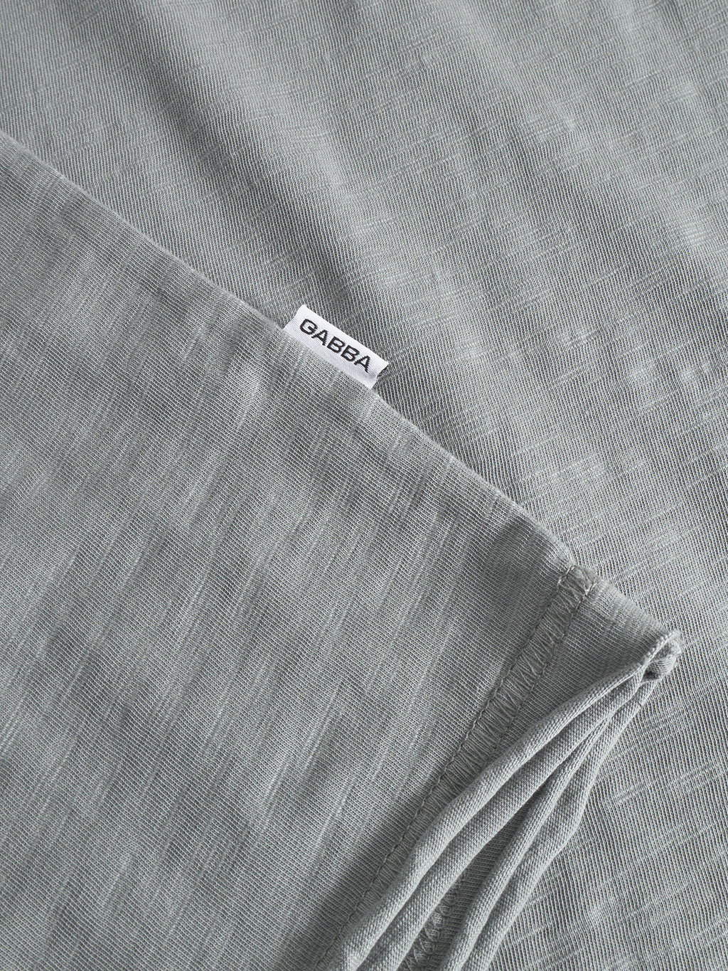 A close-up of the soft, midweight slub jersey fabric from Gabba’s Kostant New LS in Dust Sage Green, featuring a white GABBA label sewn at the edge. The material is slightly folded, perfect for a long-sleeved t-shirt.