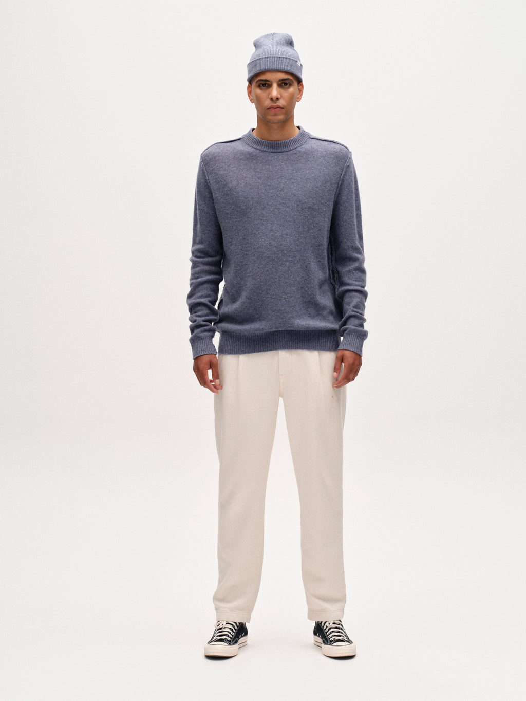 A man faces forward in a relaxed pose, wearing Gabbas Joe-Pas Vill trousers in Lt. Sand, paired with a light blue knit beanie, matching sweater, and black sneakers. The background is plain white and his expression is neutral.
