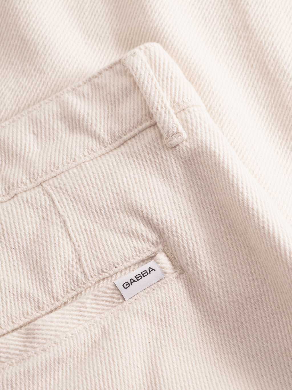 A close-up of Gabba’s Joe-Pas Vill - Lt. Sand pants in cream cotton twill shows a back pocket with the signature white label. The diagonal weave and relaxed fit make these trousers an essential wardrobe piece.