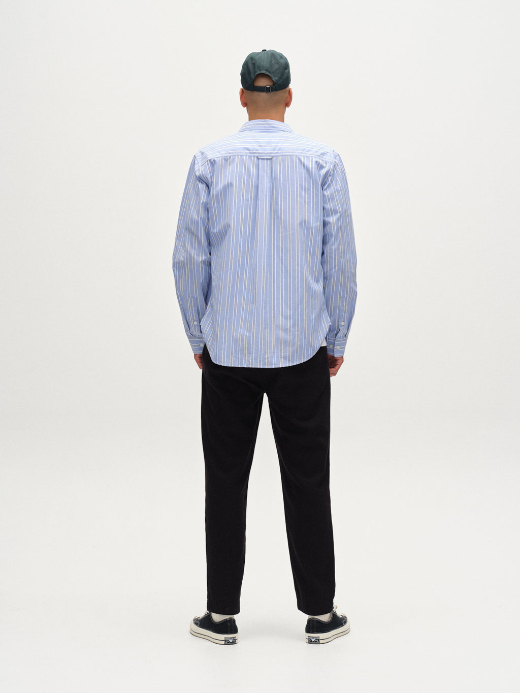A person stands with their back to the camera, wearing Gabbas Joe-Pas Vill - Black relaxed-fit trousers, a blue and white striped shirt, black sneakers, and a dark green cap against a plain white background.