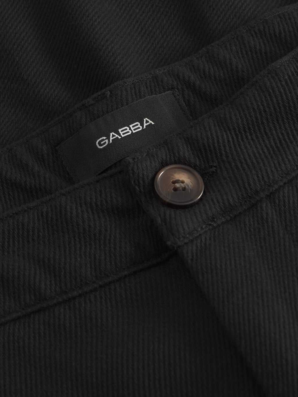 Close-up of Gabba Joe-Pas Vill - Black cotton twill pants featuring a brown button, visible black GABBA label, and subtle diagonal texture for a tailored look.