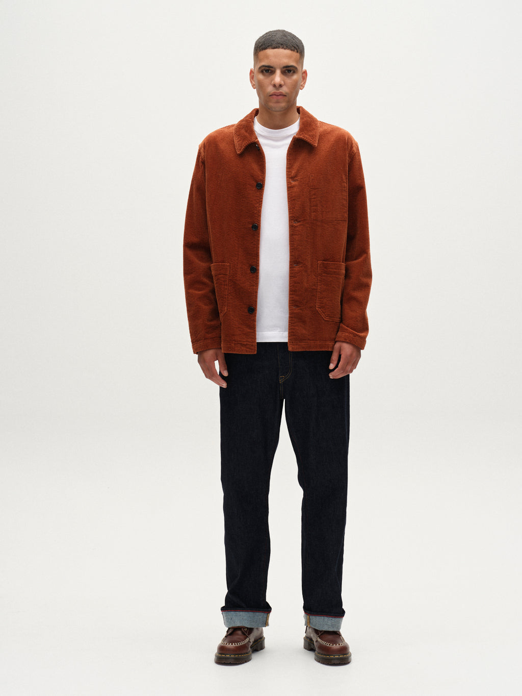 A man faces the camera with a neutral expression, wearing the Gabba Libre AX Cord in Brown Rust over a white t-shirt, paired with dark jeans and brown shoes, against a plain white background.
