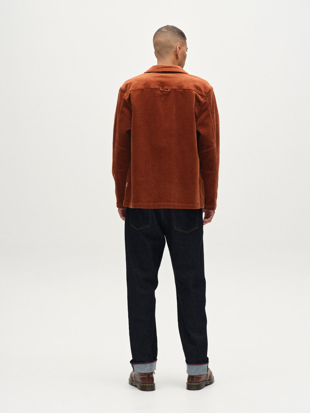 A man stands facing away in a plain, light space, wearing Gabba's Libre AX Cord overshirt in Brown Rust over a shirt, paired with dark cuffed jeans and brown shoes. His short hair is neatly styled.
