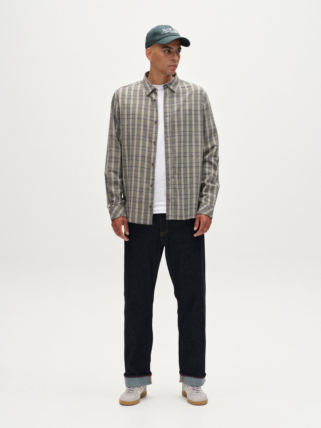 A man stands against a white background wearing the Gabba Sven Reg Herri LS - Grey check shirt over a white T-shirt, a green cap, dark jeans, and light gray sneakers. He looks to his left with his hands by his sides.