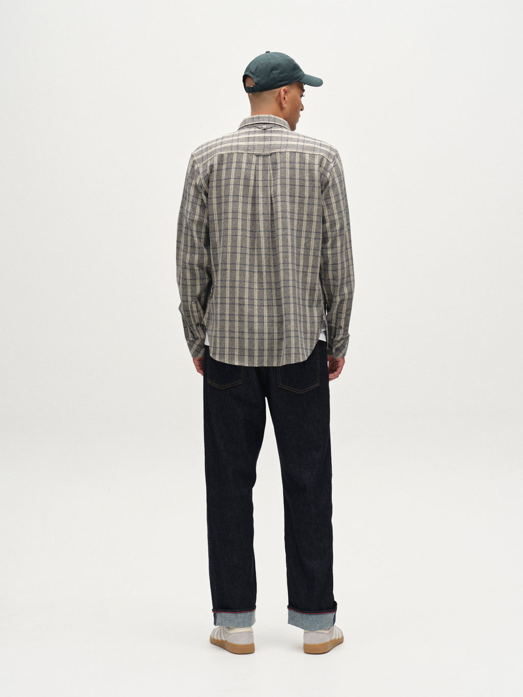 A man stands with his back to the camera, wearing a green baseball cap, dark blue jeans, white sneakers, and the Gabba Sven Reg Herri LS - Grey shirt in beige and gray plaid, against a plain white background.