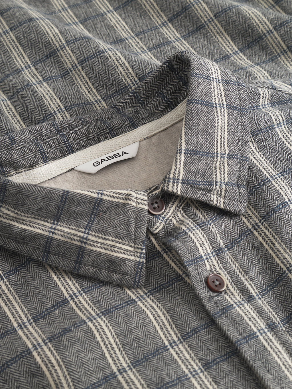 A close-up of Gabbas Sven Reg Herri LS shirt in grey and cream plaid, featuring a herringbone pattern, brown buttons, and a visible white GABBA tag on the collar.