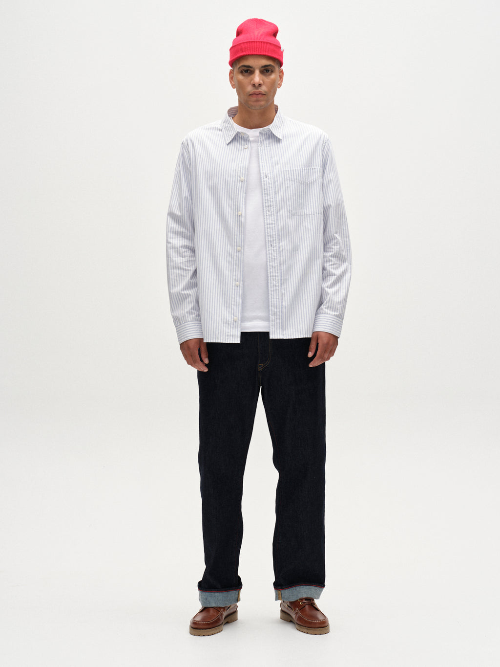 A man stands against a plain white background, wearing the Gabba Sven Reg Clo Ls - White Stripe shirt over a white t-shirt, paired with dark jeans, a red beanie, and brown leather shoes.