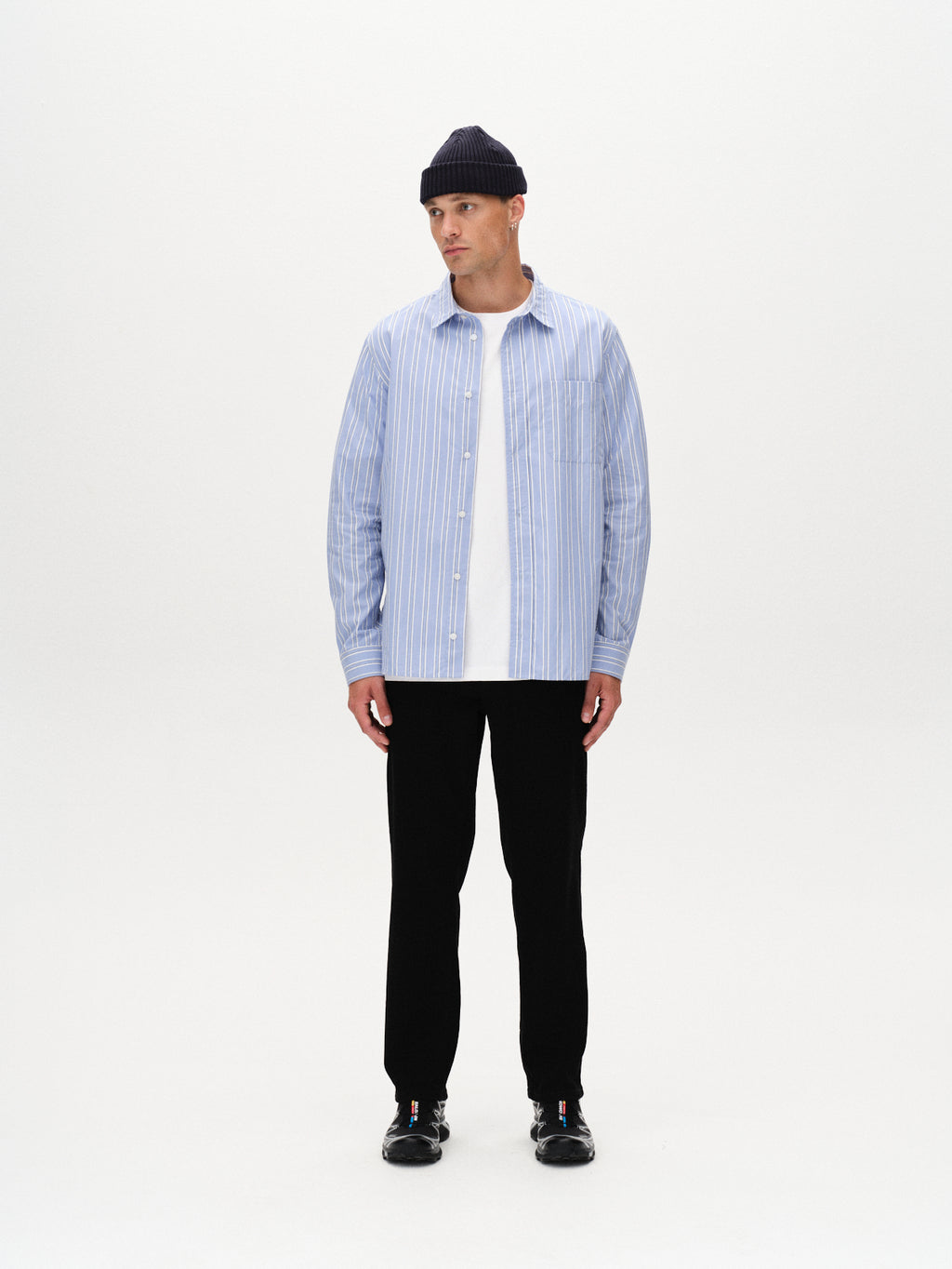 A man stands against a white background wearing the Gabba Sven Reg Clo Ls - Blue Stripe, a classic collared 100% cotton shirt, over a white tee, paired with black pants, black shoes, and a navy knit beanie. He looks slightly to the side.