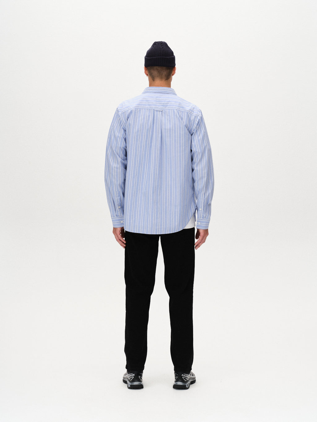 A person stands facing away from the camera wearing the Gabba Sven Reg Clo Ls - Blue Stripe shirt, paired with black pants and black sneakers, and a dark beanie, set against a plain white background.
