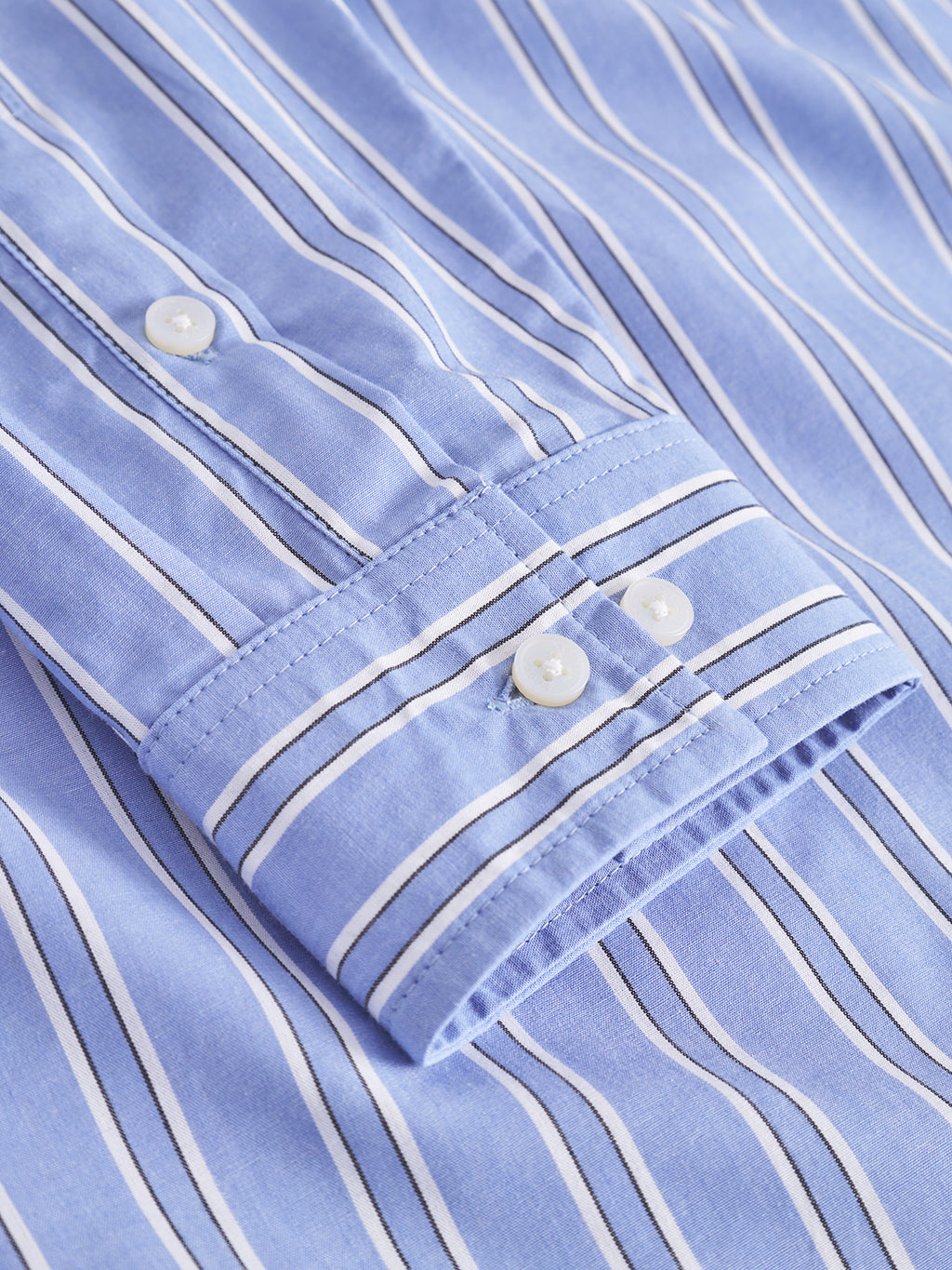 A close-up of the Gabba Sven Reg Clo Ls - Blue Stripe shirt shows 100% cotton fabric with white and navy vertical stripes, a sleeve cuff with two white buttons, and a glimpse of its classic collar design.