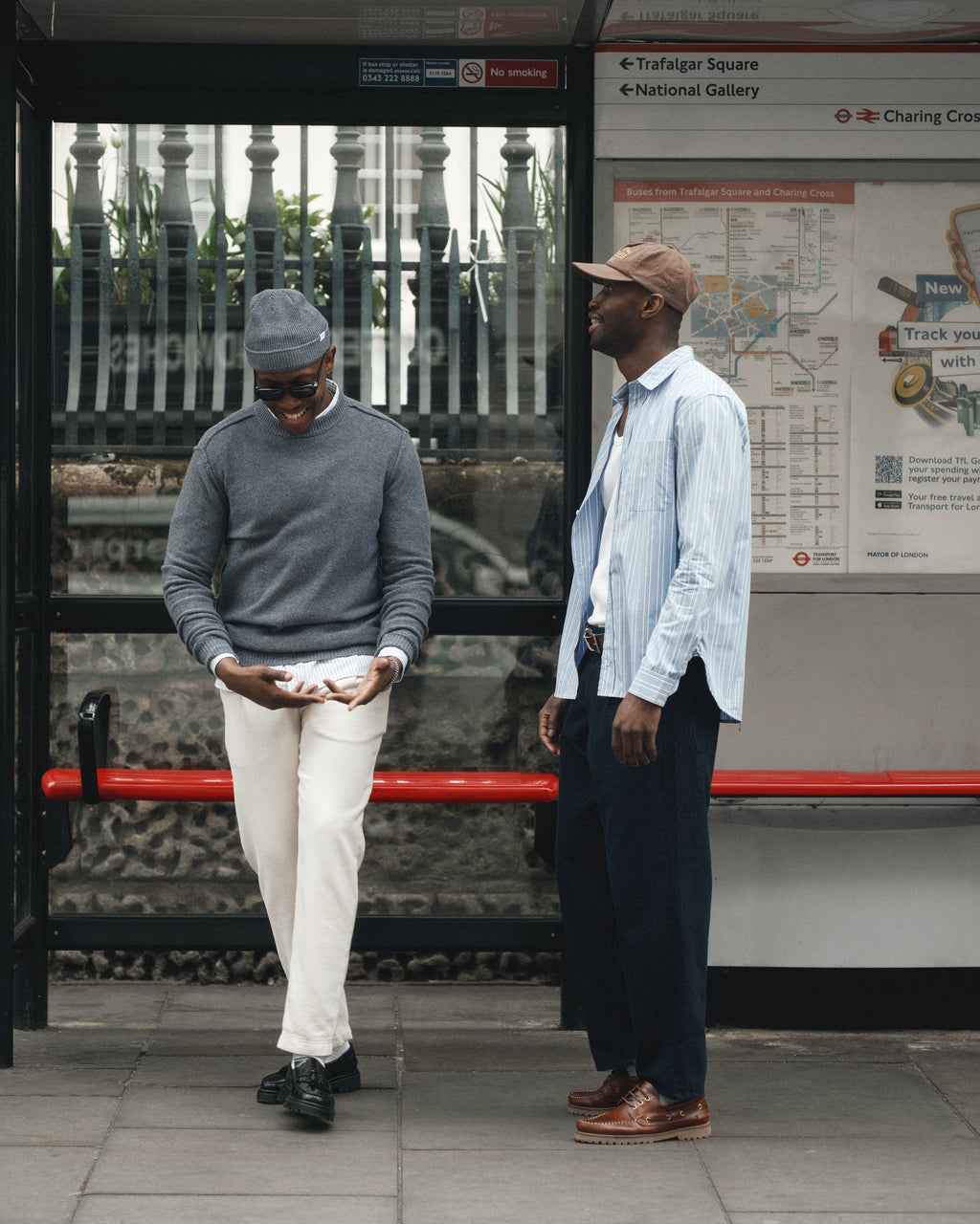 Two men stand at a bus stop. One wears a grey sweater, cap, and white pants, smiling at his phone. The other faces him in the Gabba Sven Reg Clo Ls - Blue Stripe shirt with a classic collar. A bus map hangs behind them.