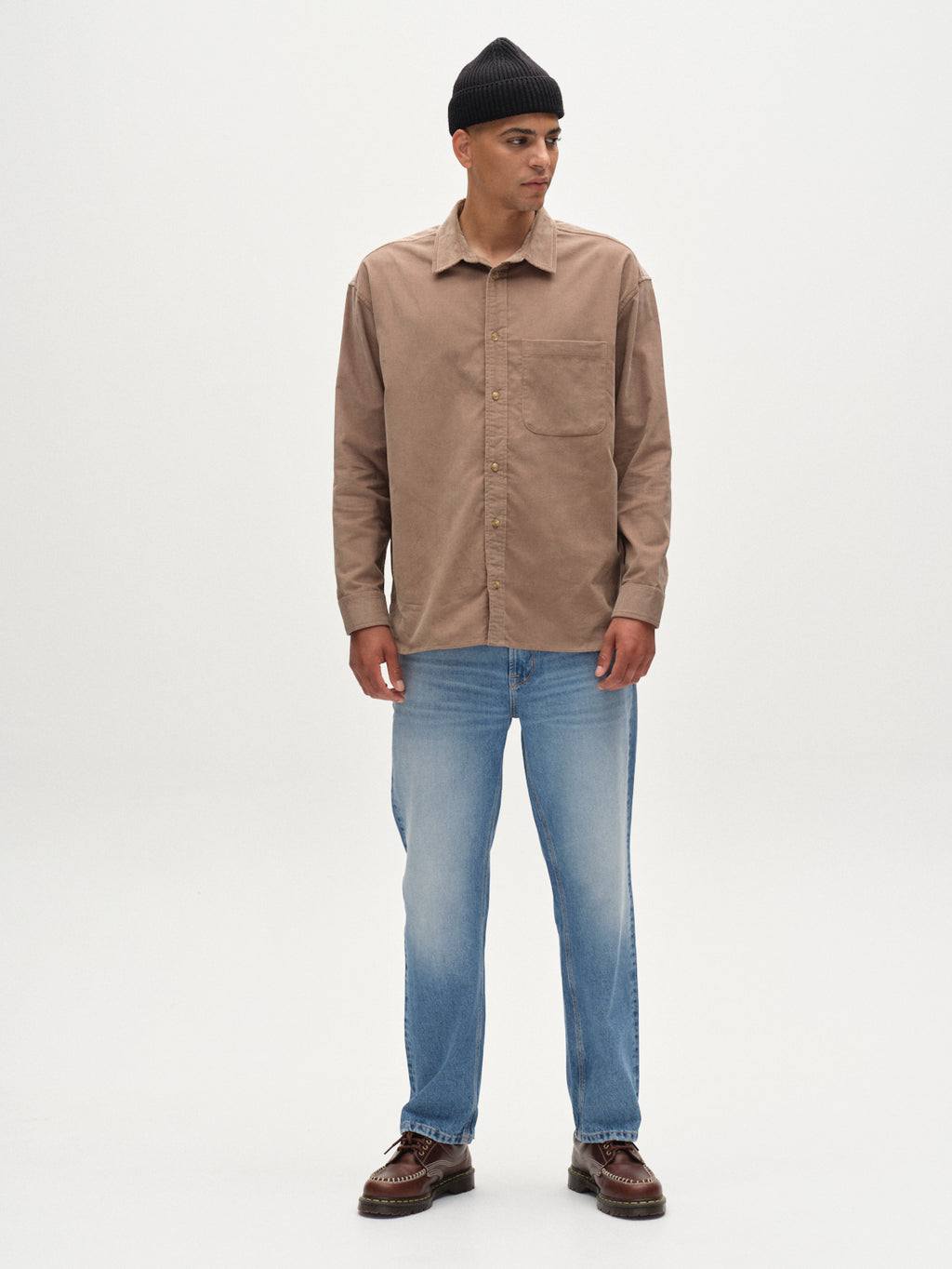 A man stands facing forward against a white background, wearing Gabbas Jens Over Cord LS - Sand Greige overshirt, a black knit beanie, light blue jeans, and brown shoes.