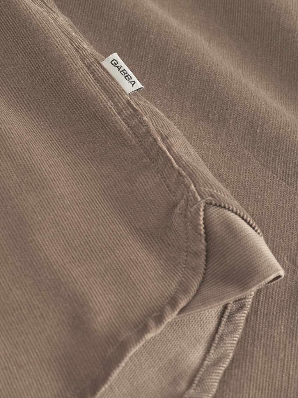 Close-up of sand greige corduroy fabric with visible stitching and a small white Gabba label, highlighting the soft texture of the Jens Over Cord LS—an ideal overshirt for cooler days.
