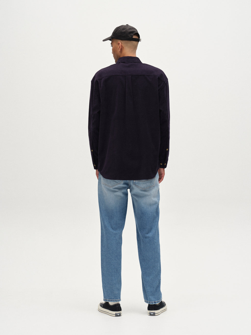 A man, seen from behind against a white background, wears the Gabba Jens Over Cord LS - Night Sky overshirt as a layering piece with a black cap, light blue jeans, and black-and-white sneakers.
