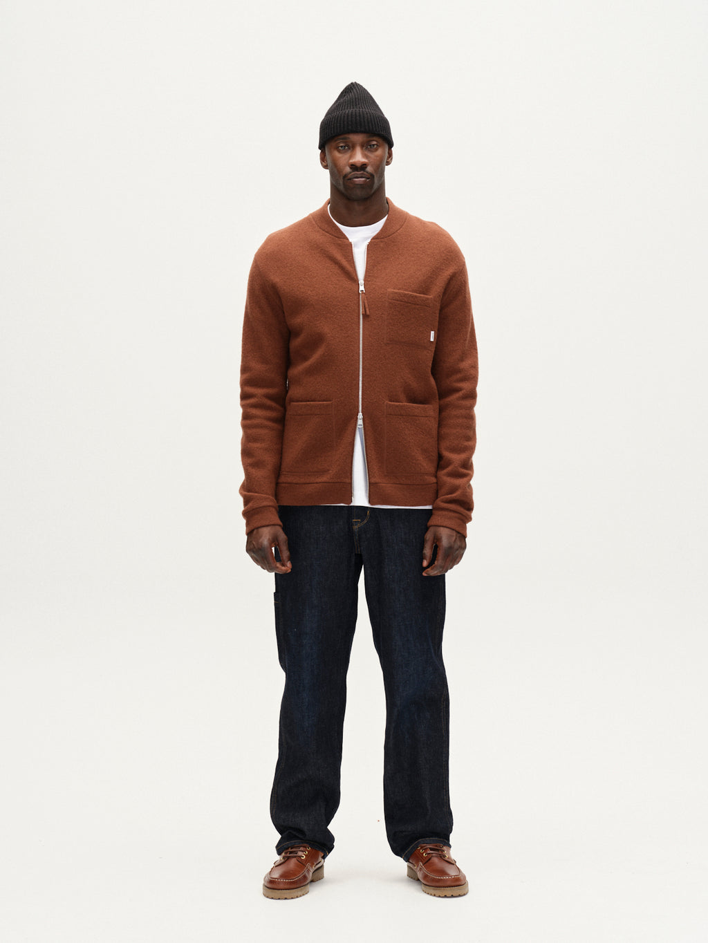 A man stands facing forward against a plain white background, wearing Gabba Max Work Self dark blue denim jeans, a black knit beanie, rust zip-up jacket over a white shirt, and brown shoes.