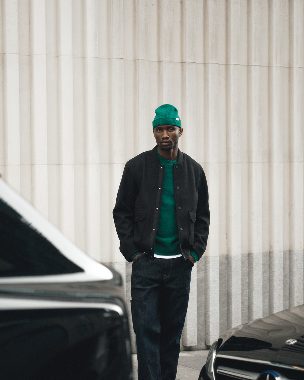A man wearing Gabbas Max Work Self - Dark Blue Denim jeans stands with hands in pockets between two black cars, set against a light-colored ribbed wall.