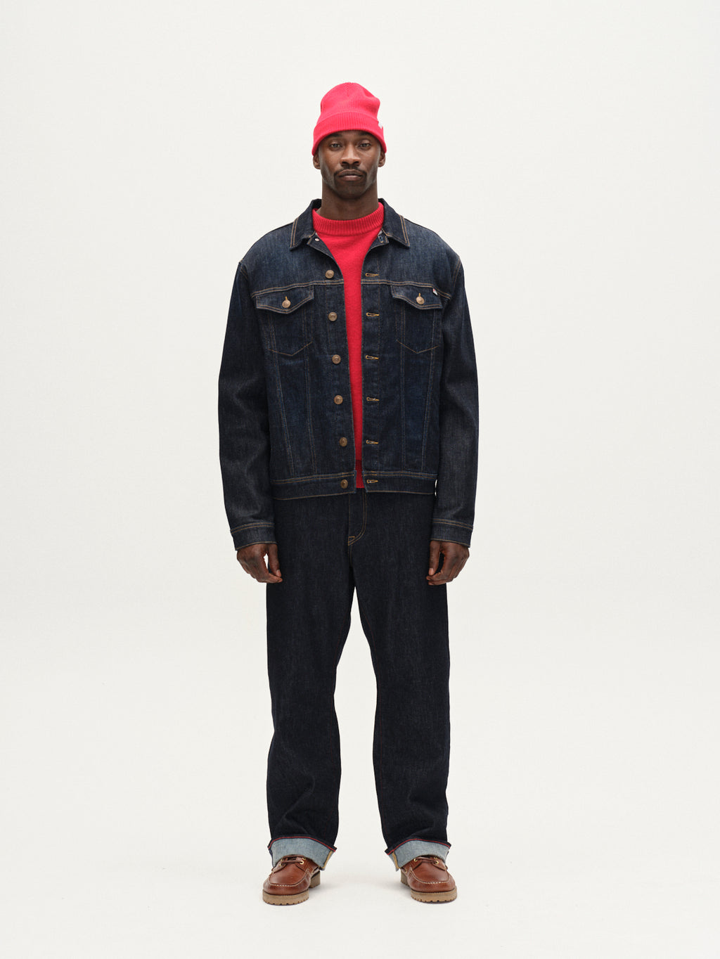 A man faces forward in a plain white setting, wearing Gabbas Luca Self - Dark Blue Denim jacket and regular-fit jeans, paired with a red sweater, red beanie, and brown shoes. His expression is neutral.