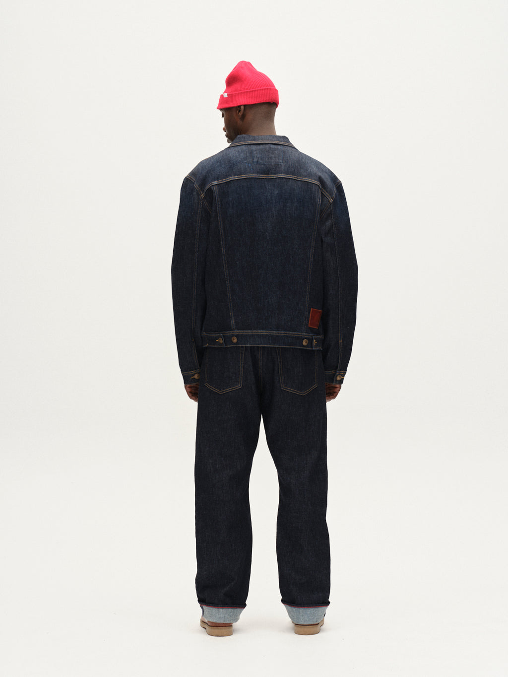 A person faces away, wearing Gabbas Luca Self dark blue denim jacket and regular-fit jeans, paired with a bright pink beanie and brown shoes, set against a plain white background.