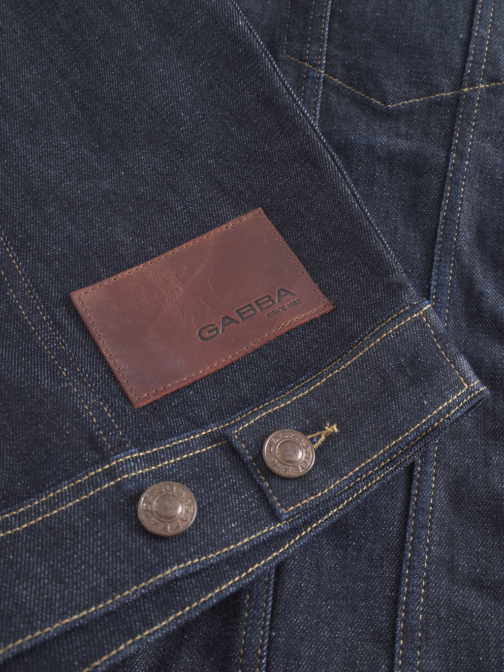 The Gabba Luca Self - Dark Blue Denim features sustainable dark blue fabric with yellow stitching, a brown leather Gabba patch, and two metallic buttons at the bottom edge.