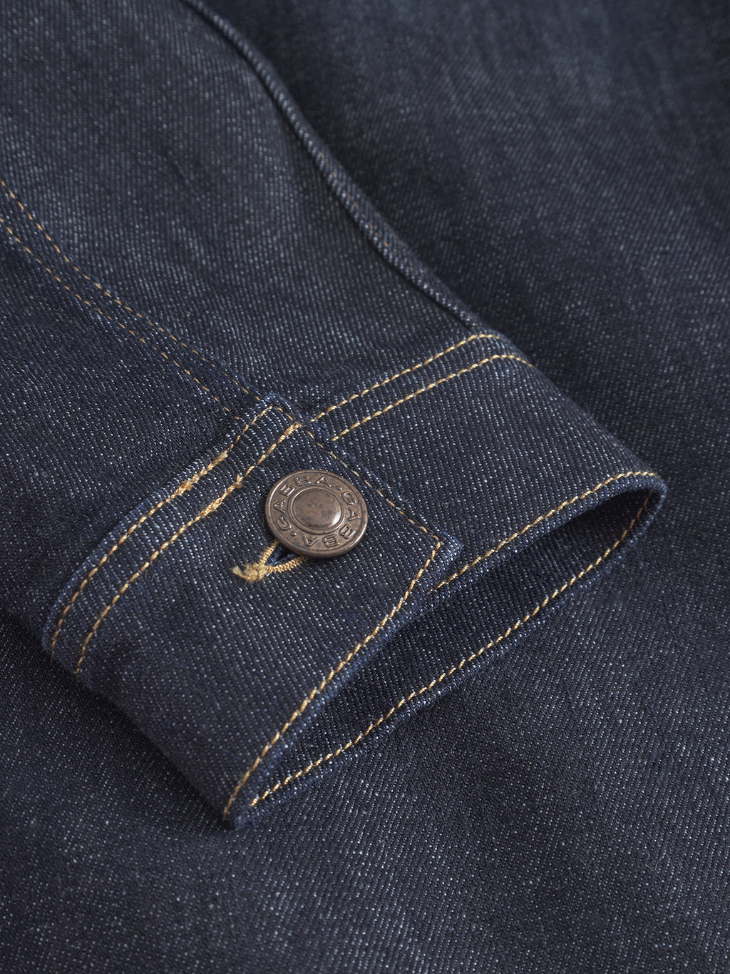 Close-up of a Luca Self - Dark Blue Denim jacket sleeve by Gabba, featuring yellow stitching and a metal button with embossed text. Made from recycled fibers, this eco-friendly piece combines style with sustainability.