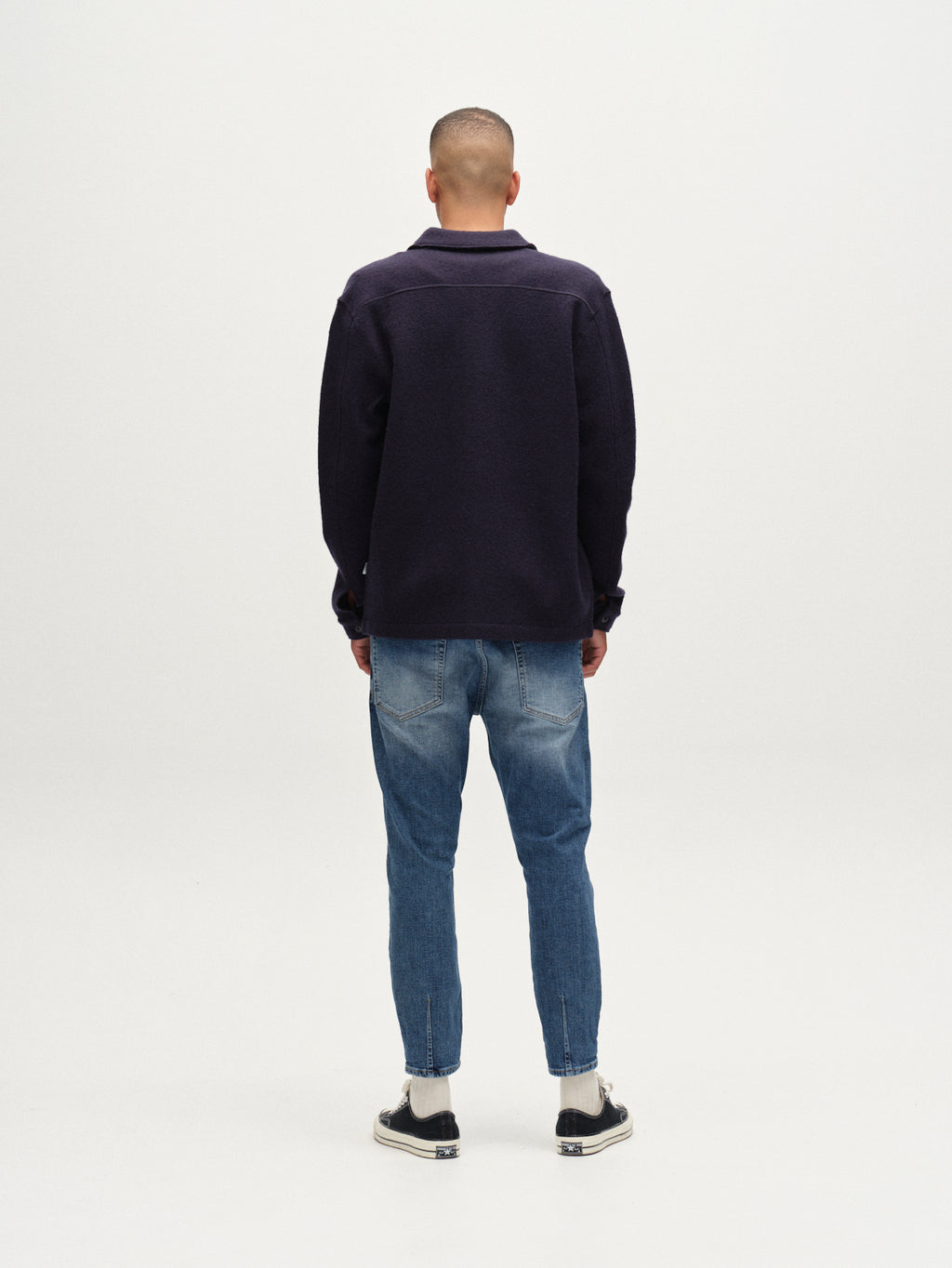 A person stands facing away from the camera in a dark long-sleeve shirt, white socks, black sneakers, and Gabbas Alex Cros - Mid Blue Denim jeans with a relaxed tapered fit against a plain white background.