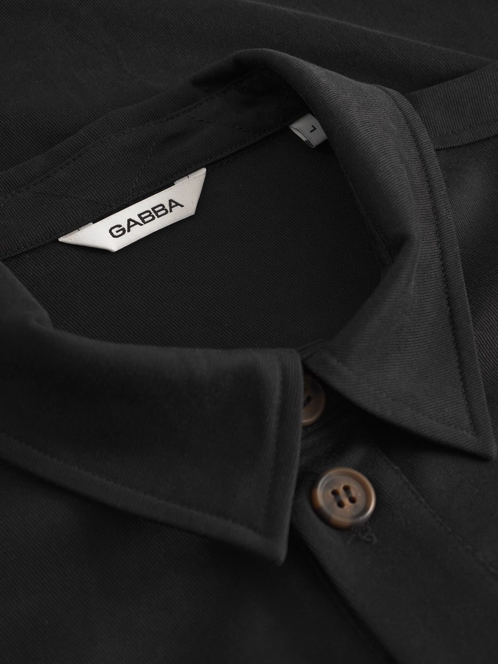 Close-up of the collar and upper buttons of the Jens Over Lupo LS - Black by Gabba, featuring brown buttons and a white brand tag—a stylish pick for layering every day.