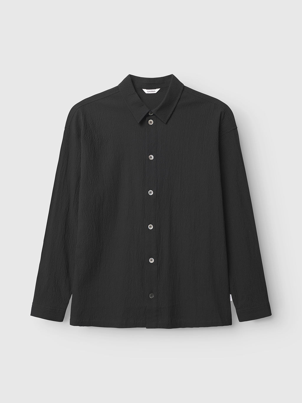 The Gabba Jens Over Siro LS GOTS - Black is a wrinkle-free, oversized long-sleeved seersucker shirt with a textured fabric, pointed collar, and white buttons, shown against a light grey background for versatile comfort and style.