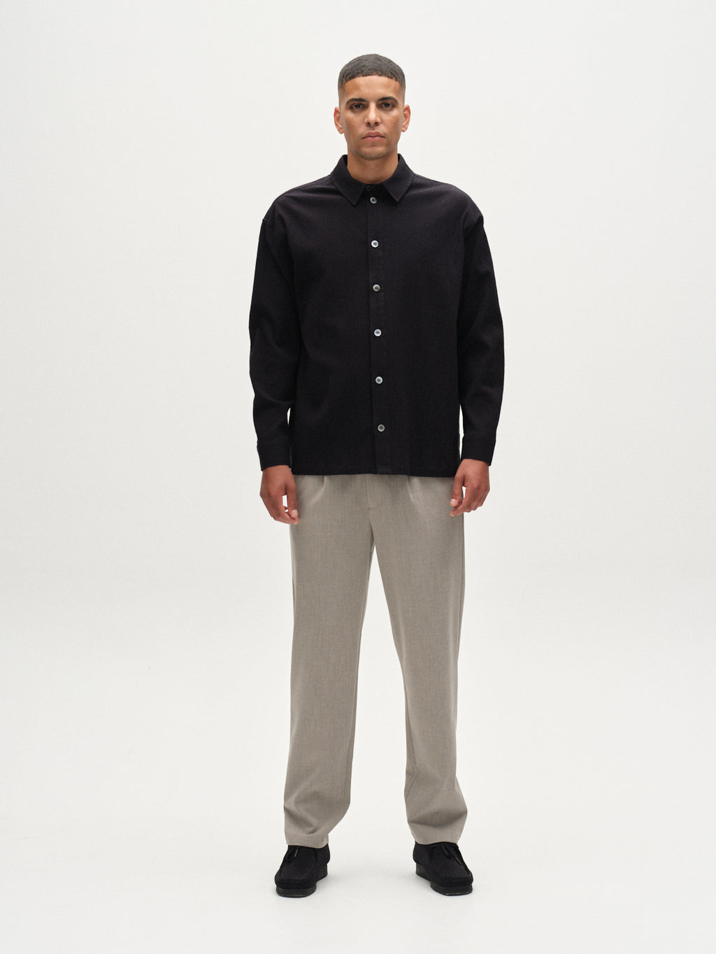 A man with short hair and a neutral expression stands facing forward against a plain white background, wearing the Gabba Jens Over Siro LS GOTS black shirt, light beige pants, and black shoes.