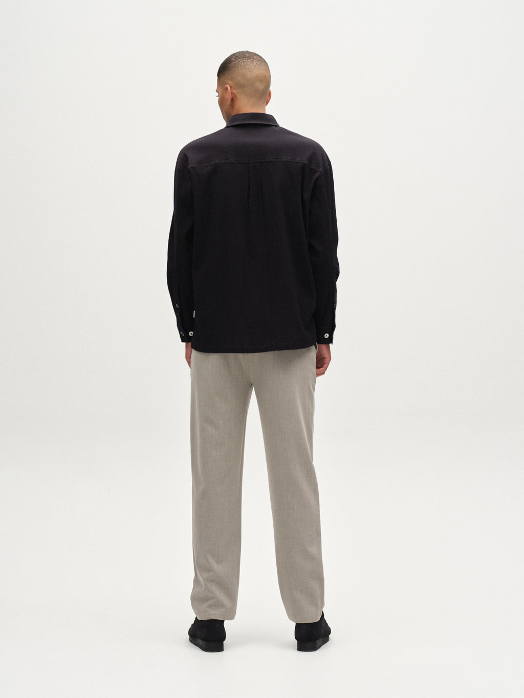 A person stands facing away from the camera, wearing the Gabba Jens Over Siro LS GOTS - Black shirt, light beige pants, and black shoes against a plain white background.