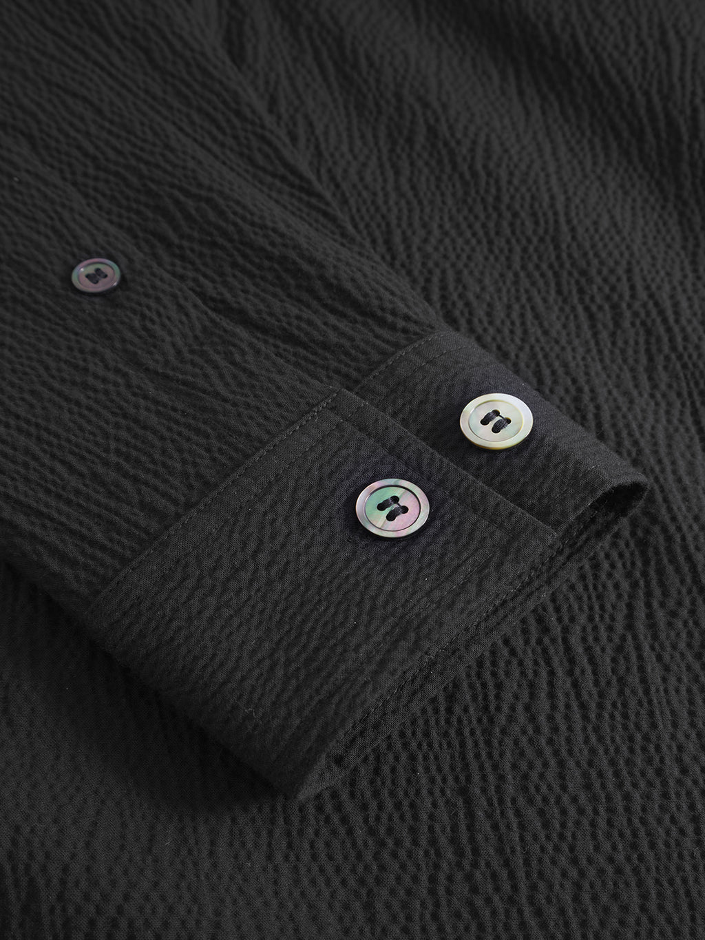A close-up of Gabba’s Jens Over Siro LS GOTS - Black shows its crinkled seersucker fabric and iridescent buttons on the cuff and sleeve—ideal for anyone seeking an oversized black shirt with unique texture.