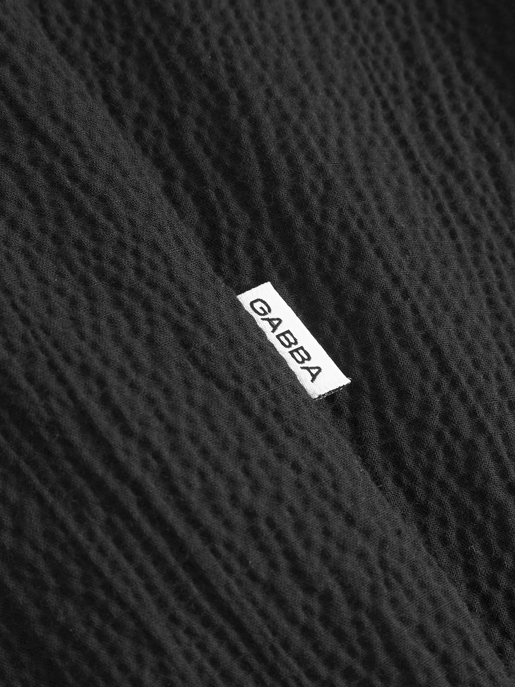 Close-up of textured black fabric from the Gabba Jens Over Siro LS GOTS shirt in black, featuring a small white Gabba label stitched onto the material.