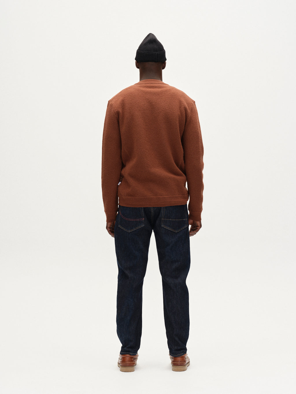 A man stands facing away from the camera, wearing a brown sweater, Gabba Carl Anark Self dark blue denim jeans, brown shoes, and a black knit beanie against a plain white background.