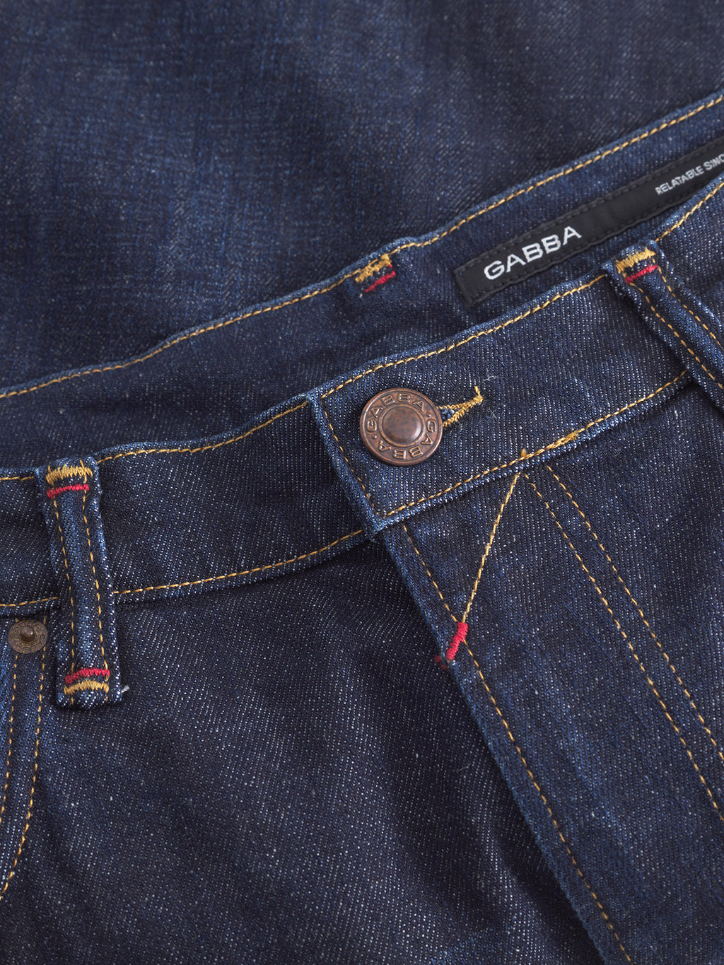 Close-up of Gabba’s Carl Anark Self dark blue denim jeans with yellow stitching, metal button, and visible waistband label. Features Japanese-inspired details and vivid red and yellow thread accents on belt loops and pocket seam.