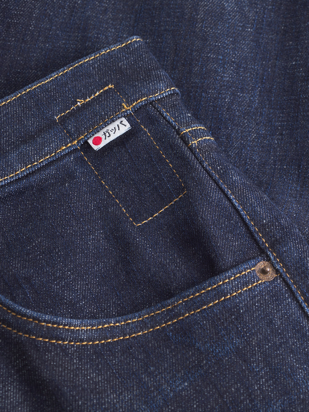 A close-up of the Carl Anark Self - Dark Blue Denim by Gabba shows front and coin pockets with a small fabric tag featuring Japanese text and a red dot. Yellow stitching highlights the seams on these dark blue loose tapered jeans.