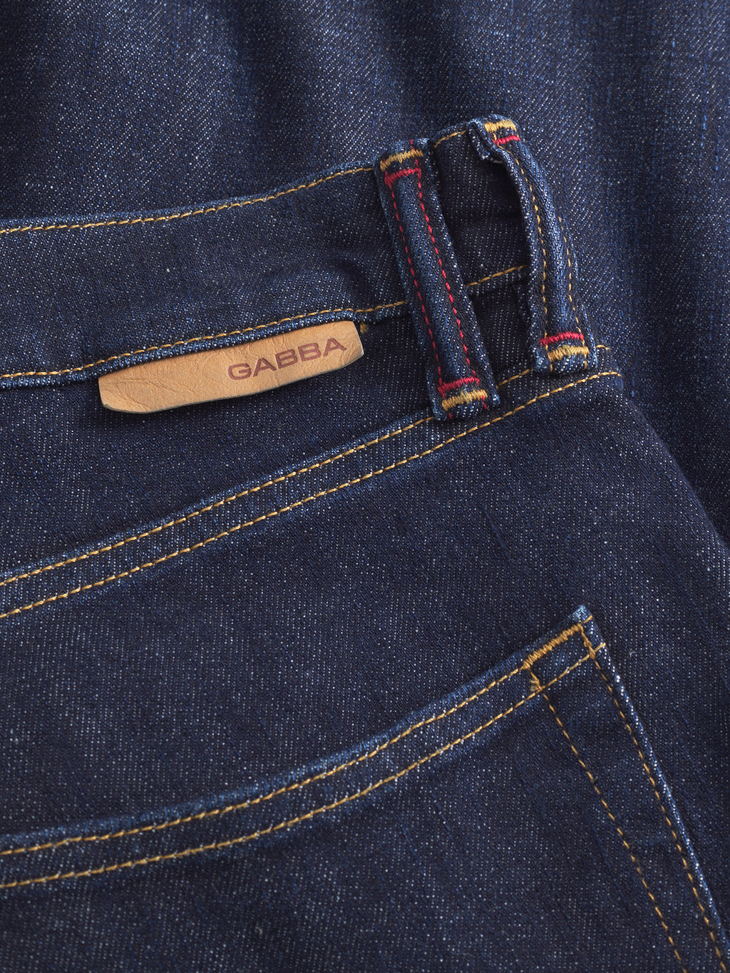 Close-up of Gabbas Carl Anark Self dark blue denim jeans, featuring yellow stitching, a leather branded patch, and belt loops with red and yellow thread accents.
