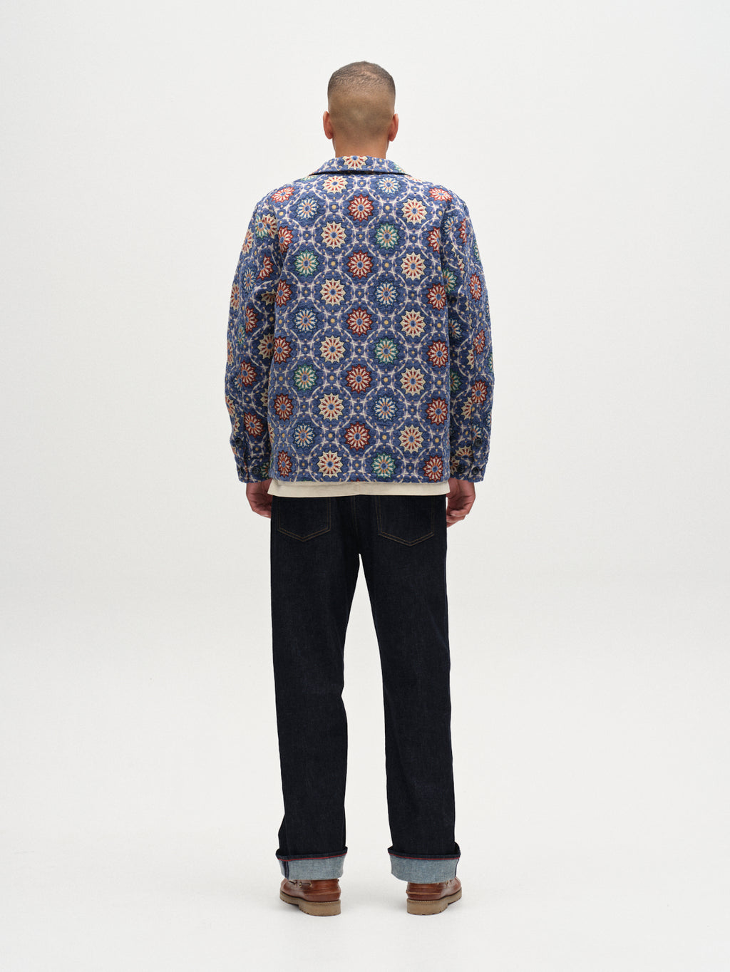 A person stands facing away, wearing the Gabba Simone Nava LS - Multi Pattern jacket with colorful designs, paired with dark jeans and brown shoes, set against a plain white background.