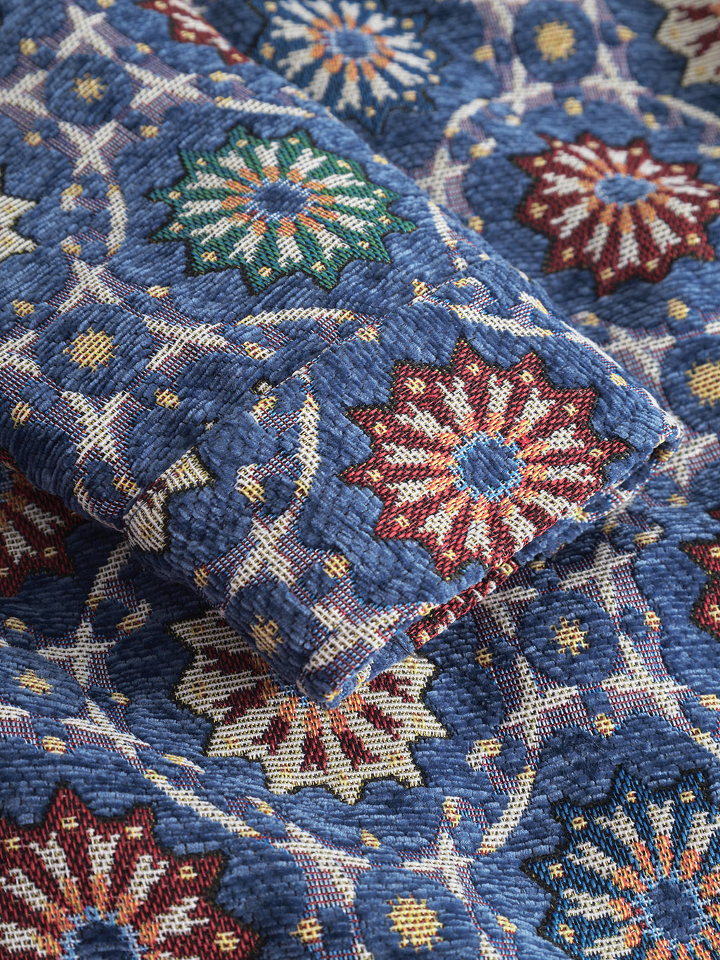 A close-up of Gabbas Simone Nava LS - Multi Pattern textile shows thick, slightly rough blue jacquard brocade woven with intricate, colorful floral star designs in red, green, and yellow.