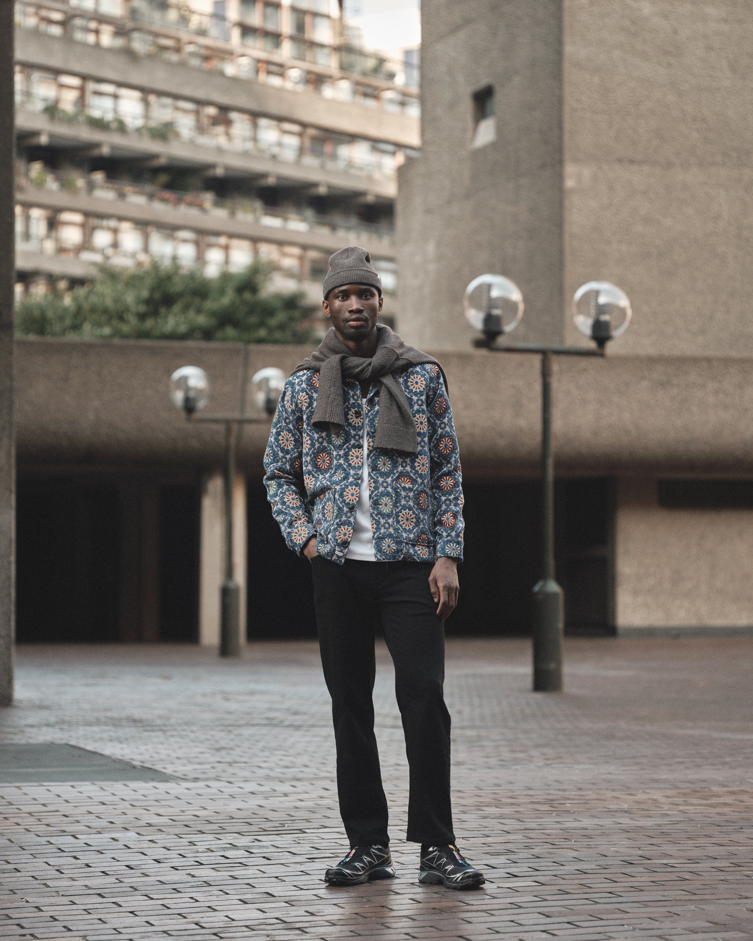 A man stands on a brick path before a concrete building, wearing the Gabba GABsimone Nava LS - Multi Pattern shirt, black pants, sneakers, a brown knit hat, and a sweater draped over his shoulders.