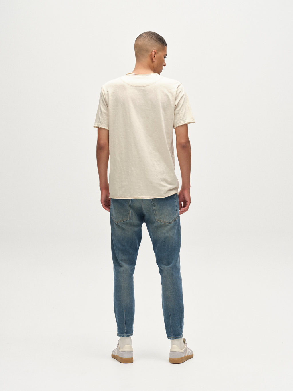 A man faces away from the camera in a cream T-shirt, Gabbas Alex Ulio - Mid Blue Denim relaxed tapered jeans with stretch comfort, and beige sneakers, set against a plain white background.