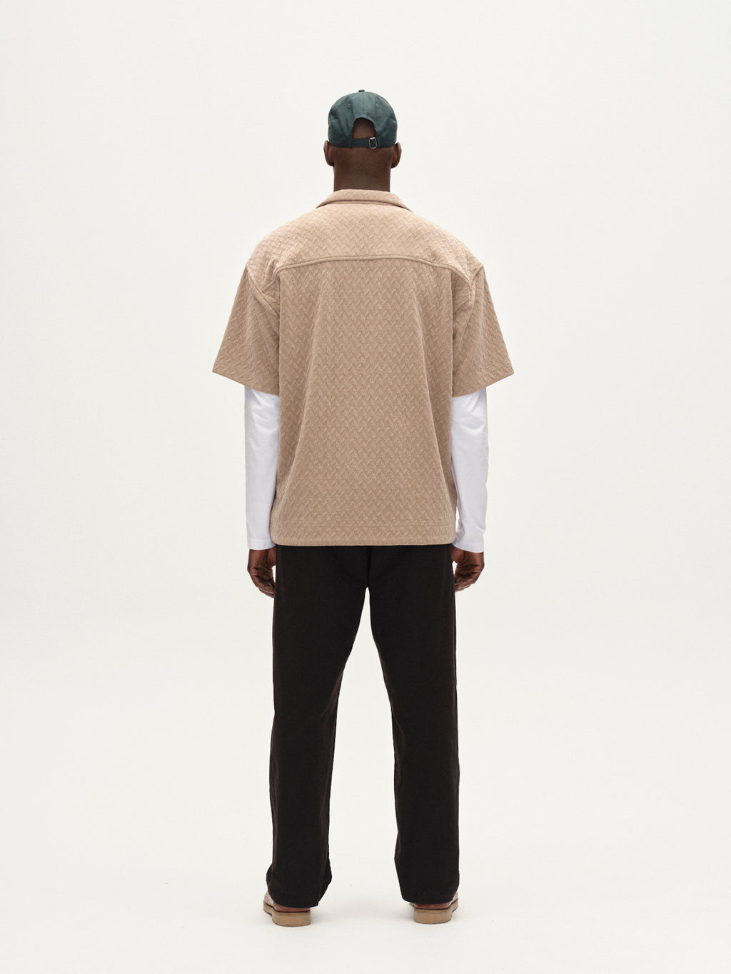 A person stands facing away from the camera, wearing the Gabba Jens Over Diam SS shirt in Sand over a white long-sleeve top, paired with a green cap, dark pants, and beige shoes against a plain white background.