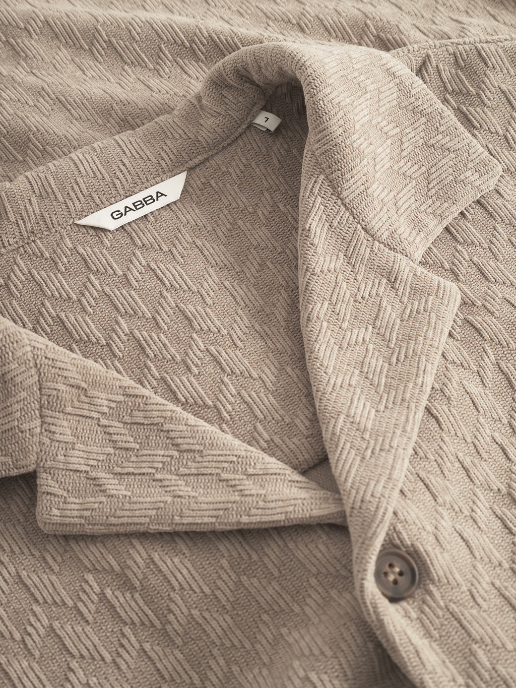 Close-up of the Jens Over Diam SS overshirt by Gabba in sand—a textured beige jacquard velvet jacket with patterned fabric, visible collar, button, and an inner white Gabba label, perfect for refined and elevated looks.